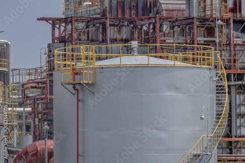 Foto Close-up of large industrial storage tank with red fire suppression pipe and yellow safety railings, backed by maze of rusted pipes and steel frameworks of an aging refinery