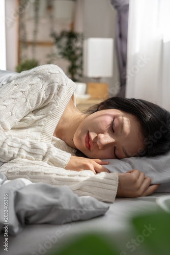 Young Asian woman sleeping on comfortable bed in morning sunlight looking peaceful and relaxed resting head on soft pillow wearing cozy white sweater enjoying quiet nap. Vertical