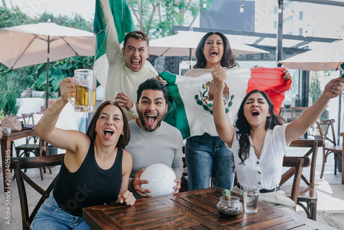 latin people soccer fans watching and cheering a match game with goal, victory or winning championship in a terrace pub. Group of mexicans friends holding a flag in celebration at bar together