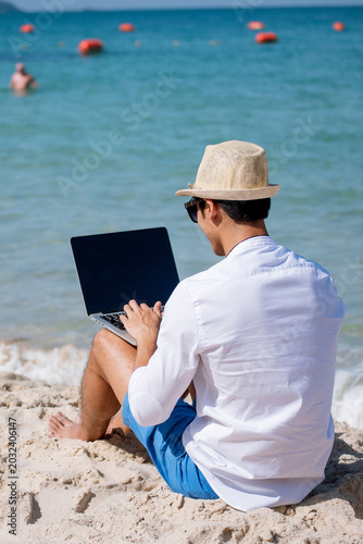 Vertical Man traveler use laptop on summer beach blue sky. Man legs walking on tropical beach. Relaxation Travel footstep. Asian young men wear casual short pants tropical smiling face