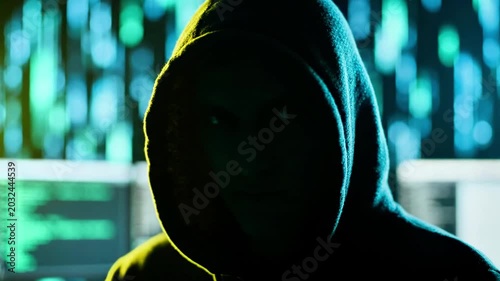 A hooded figure gazes intently in a dimly lit room with neon-lit computer screens