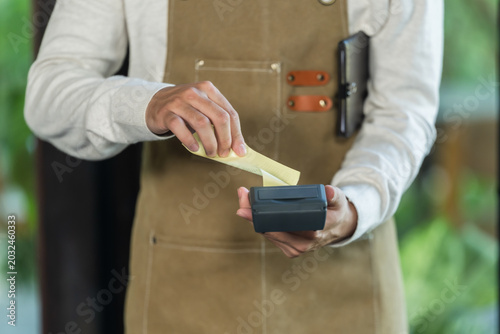 Asian son pulls printed receipt from payment terminal in family cafe, finalizing billing proof, cashless checkout record, front counter routine, restaurant transaction workflow training.
