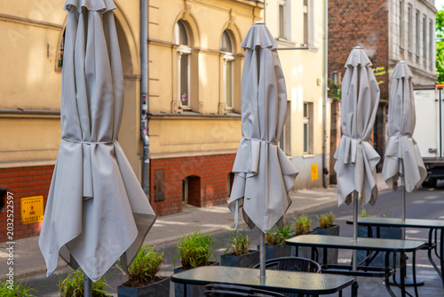 Bild auf Leinwand An outdoor restaurant patio on a city sidewalk featuring a row of closed light grey umbrellas, glass tables, and black chairs against historic tenement buildings with yellow and brick facades