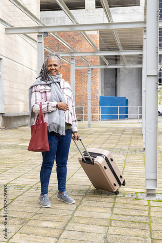 Senior African American woman standing under canopy in jacket holding beige suitcase and red tote