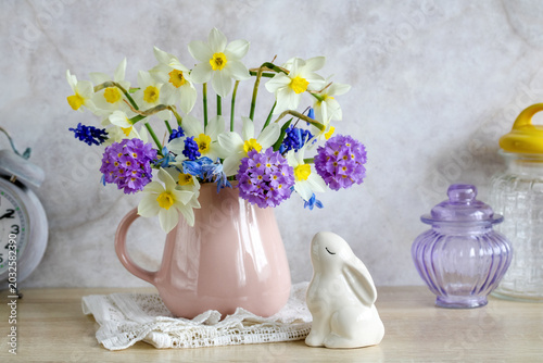 Easter still life, bouquet of garden flowers of daffodils, primroses and mouse hyacinths in a jug and a rabbit figurine