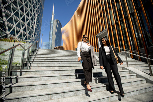 Fotografi Women walk down steps in a busy urban area during bright daylight hours