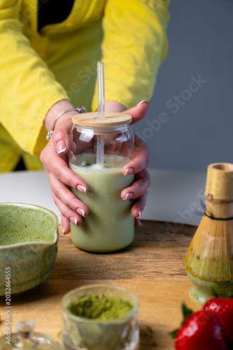 Portable matcha latte in glass jar on tray with ceramic bowl and fresh strawberries in morning light. Hands near chilled green drink ready for commute. On the go matcha, portable wellness drink, busy