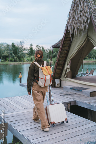 Eco-tourism and safe travel insurance concept Asian woman traveler with backpack pulling suitcase arriving at beautiful lake glamping tent enjoying peace of mind during nature weekend holiday vacation