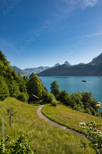 The path to the Rütli meadow in summer with a blue sky, Lake Lucerne with a boat sailing on it, and the Mythen mountain.