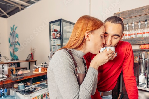 red-haired woman and a young man share a moment at a cafe. The woman sips coffee while the man leans in, smiling. The cafe has a modern decor.