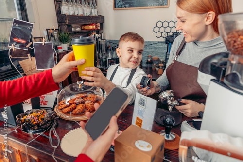 smiling mother and little boy handing takeaway coffee cup to customer in modern coffee shop interior while working together behind counter. family business, cafe service and coffee culture