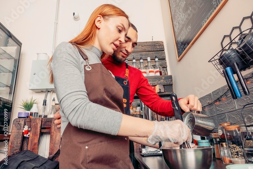 A young Caucasian woman and a young Middle-Eastern man prepare coffee in a cozy cafe. They are focused on their task, surrounded by coffee-making equipment.