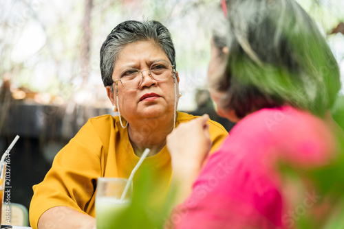 Senior Asian woman in yellow top listens closely during serious reunion talk at garden cafe, hearing painful update from friend in pink, offering empathy, patience, care, guidance, trust, comfort.