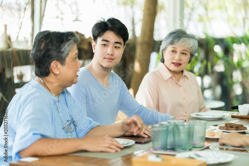 Senior Asian woman in blue blouse talks across cafe table with adult son and elderly relative, sharing update, advice, and caring reunion conversation before meal in bright garden restaurant.