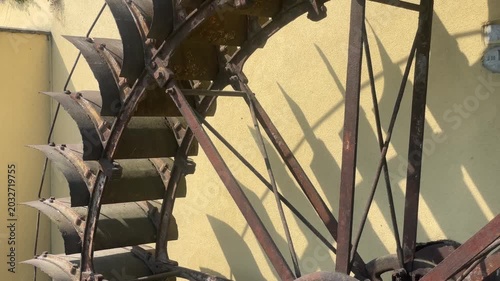 Rustic Water Wheel: A weathered, industrial water wheel made of metal and wood stands against a yellow wall, casting long shadows under the bright sun. Robbiano, Italy