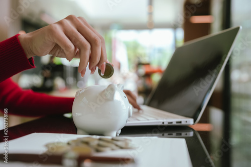 Close-up of a woman putting coins into a white piggy bank, representing saving money, personal finance, budgeting, and future financial security.