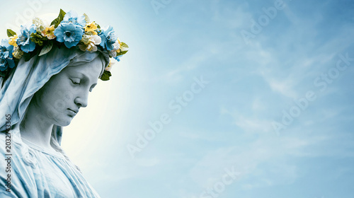 Beautiful marble statue of the Virgin Mary wearing a vibrant flower crown, looking down in a serene and prayerful pose against a clear blue sky background with ample copy space for text.