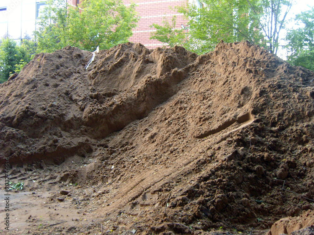 Fototapeta premium A large pile of excavated brown soil sits at a construction site. Heavy machinery tracks mark the ground at its base. In the background, green trees and a red brick building are visible under daylight