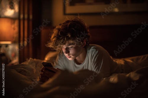 Teenager alone in bed scrolling on phone in dim light, feeling isolated