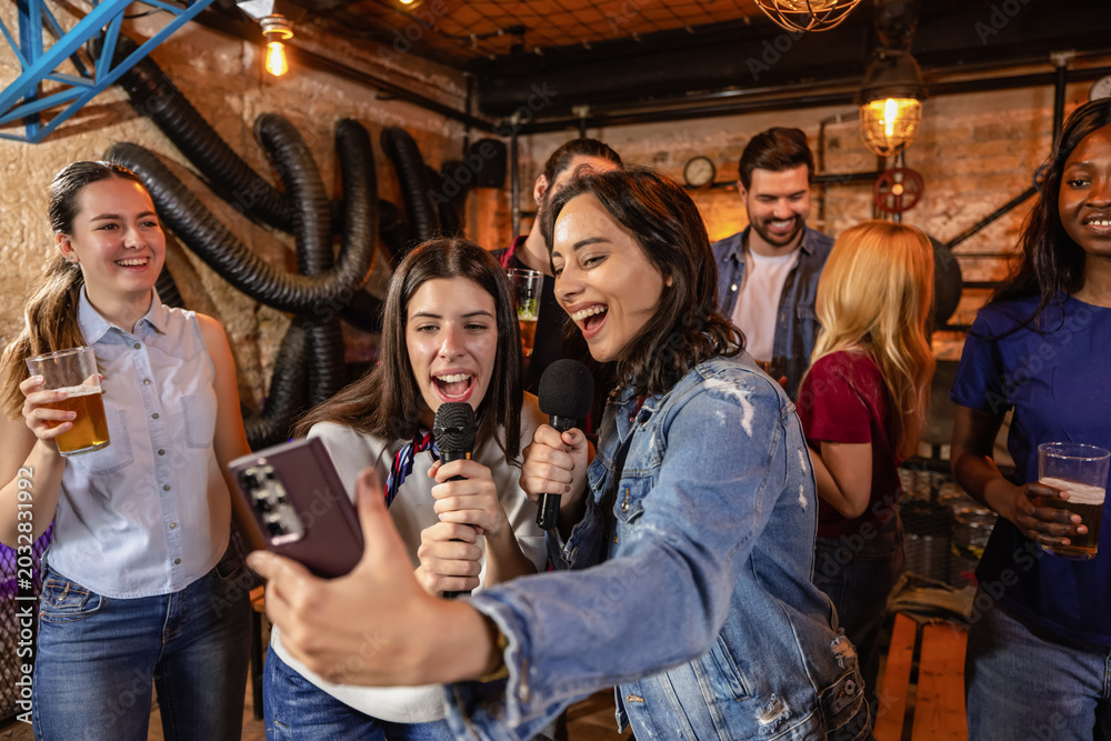 Fototapeta premium Friends singing karaoke in modern pub with beer and microphone