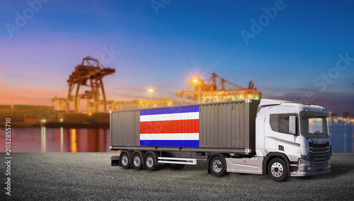 Photos Costa Rica Shipping Container Truck: A modern truck carries a shipping container emblazoned with the Costa Rican flag, ready for transport at a bustling port with cranes and twilight sky