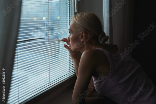 Konstfotografi A blonde woman looks out of a window through white horizontal blinds in a dimly