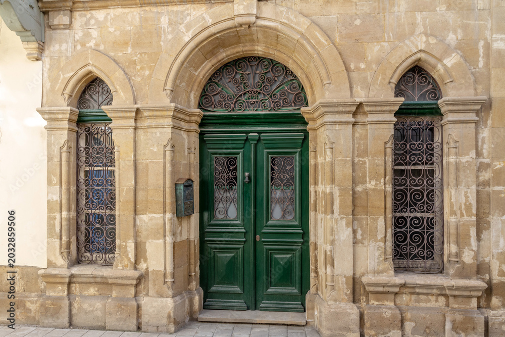 Fototapeta premium Vintage building facade made of limestone blocks with green wooden door. Traditional mediterranean architecture with arched portal and iron forged grates on windows.