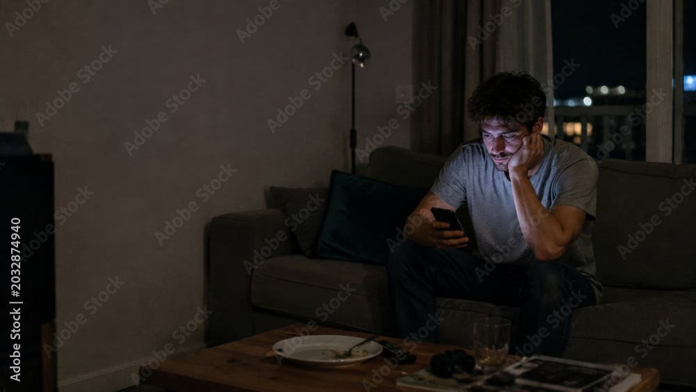 Obraz premium Man sitting on sofa at night looking at smartphone, feeling lonely in dimly lit living room