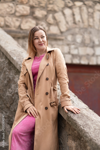 Fotografi Woman Trenchcoat Fashion: Woman in coat poses on stone wall, smiling during daytime for fall style