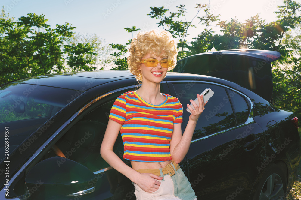 Fototapeta premium Smiling woman with curly hair wearing a striped croptop stands beside a sleek car on a sunny day, expressing a vibrant lifestyle and retro fashion mood. Soft daylight enhances warm tones, suggesting