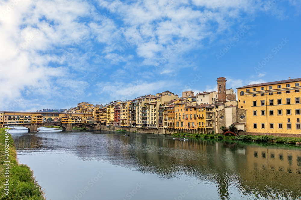 Fototapeta premium Ponte Vecchio bridge stands over the Arno River in Florence. Surrounding buildings reflect the rich history of the city. The sky is clear and the water flows gently.