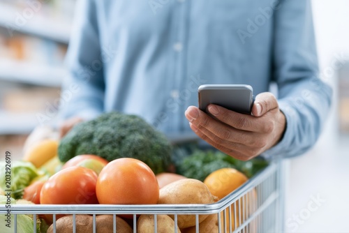 Man holding shopping cart with groceries and smartphone, smart shopping, grocery store, digital coupons