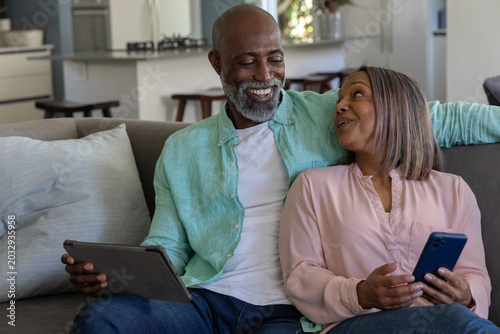 Couple sitting on gray couch in home living area holding tablet and blue smartphone