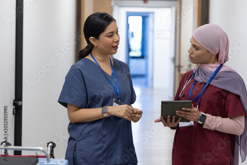 Two female medical workers in scrubs standing in hospital corridor discussing tablet, stethoscope