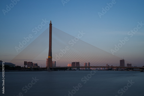 The iconic Rama VIII Bridge in Bangkok spans the Chao Phraya River at dusk. Its sleek asymmetric pylon and cables stand against a soft gradient sky while city buildings glow in the fading light.