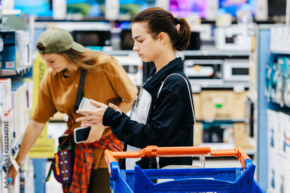 Fototapeta premium Two young women shopping for electronics in a retail store aisle, one checking a product while the other browses nearby with a shopping cart