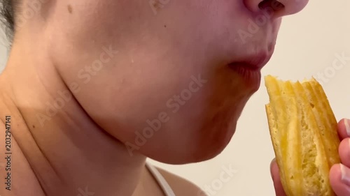 A fair-skinned woman takes a bite of a sweet eclair, delicately holding it between her fingers, showing off its golden texture and appetizing appearance in a well-lit room.
