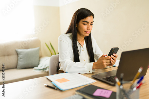 Hispanic business attorney remotely working from home, reading messages on smartphone while using laptop at office desk