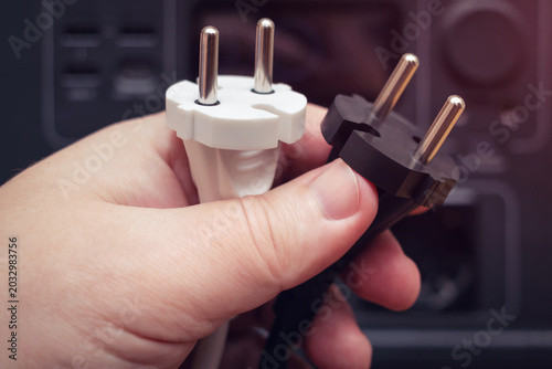 Hand holding black and white European electrical plugs at out of focus power equipment background.