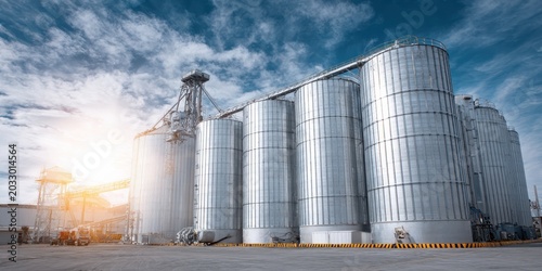 Industrial farm agricultural silos for storage and drying of wheat grains, sugar, corn, soy, sunflower on blue sky. Large metal grain tanks processing equipment in modern facility. Agriculture, food.