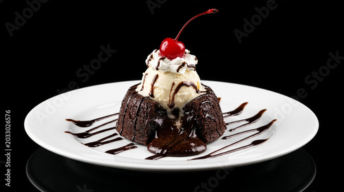 Quadro su tela Decadent molten chocolate lava cake topped with vanilla ice cream, whipped cream, and a bright cherry, served on a white plate against a black background