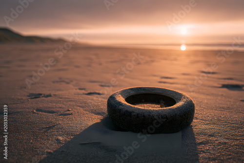Old tire on a desolate sandy beach at sunset, bathed in golden light. A stark reminder of pollution's impact on natural beauty.