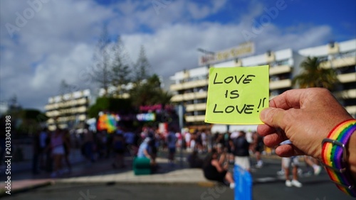 Person wearing a rainbow wristband holds a yellow sticky note with LOVE IS LOVE!! written on it, expressing support for LGBT rights and equality during a Pride event