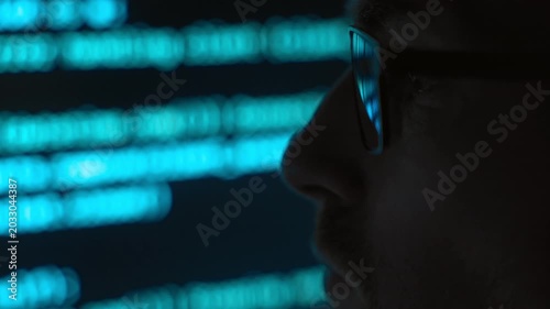 Side view close up, focused software engineer in eyeglasses looking at a blue code running on screen in a dark room, programming script reflects in his lenses. AI and data science concept. Copy space