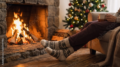 Person wearing cozy wool socks relaxes in a comfortable armchair while holding a warm mug near a glowing stone fireplace
