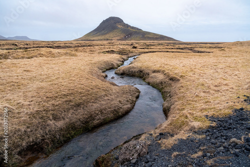 River flows through a dry grassland with a mountain in the background on the Reykjanes Peninsula in Iceland during a cold March day.