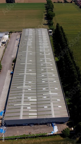 Industrial Warehouse Rooftop Aerial View: An expansive industrial warehouse with a corrugated metal roof is viewed from above, surrounded by agricultural fields and lined with trees.