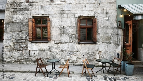 Rustic stone building facade with outdoor cafe seating in historic street. Empty cafe tables and chairs beside an aged stone wall in a picturesque old town setting