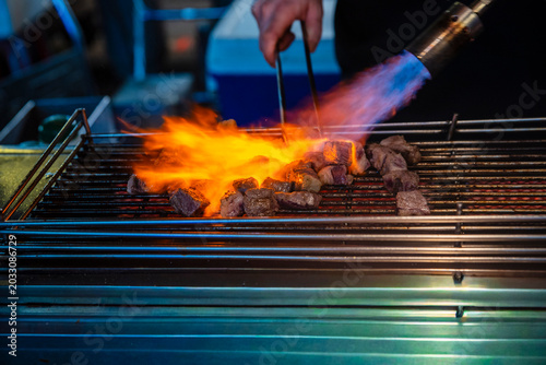 Foto A chef using a gas blowtorch to flame-grill beef cube steaks on a wire rack at a night market