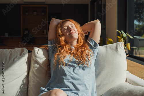 Quadro su tela Young adult woman with red hair wearing pajamas comfortably relaxing on a living